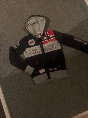 Geographical Norway hoodie med tryck - Cool hoodie från Geographical Norway med huva, dragkedja och flera broderade och tryckta detaljer. Färgerna är mörkblå, grå och röd med norska flaggan på bröstet. Materialet är mjukt och bekvämt, perfekt för chill dagar. Snygga detaljer på ärmar och rygg.