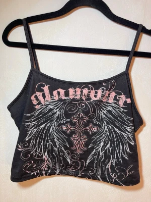Svart croppad linnetopp med tryck - Snyggt svart croppat linne med tunna axelband och tryck i rosa och vitt. Framsidan har ett stort motiv med vingar, kors och texten 'glamour' i gotisk stil. Perfekt för dig som gillar edgy och unik stil.  Fint skick, inga skavanker. SHEIN, storlek M. Djur, rök och parfymfritt hem.