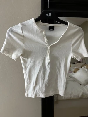 Vit ribbad topp från Gina Tricot - Säljer en vit ribbad topp från Gina Tricot med korta ärmar och knappar framtill. Toppen har en croppad passform och är tillverkad i mjuk bomullsmix. Perfekt till jeans eller kjol för en enkel och fräsch look.