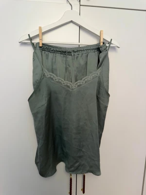 Grågrön satinpyjamas med spets - Snygg pyjamas i grågrön satin med tunna axelband och spetsdetalj vid urringningen. Setet består av ett linne och shorts med resår i midjan. Perfekt för mysiga kvällar hemma.