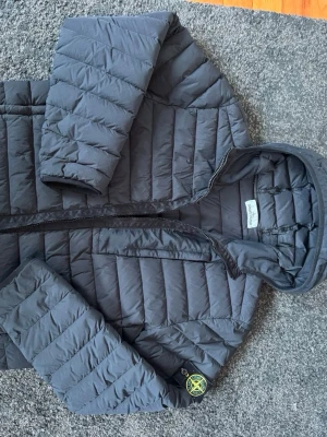 Svart pufferjacka från Stone Island - Säljer en svart pufferjacka från Stone Island med klassisk patch på ärmen. Jackan har huva, dragkedja framtill och horisontella sömmar som ger en schysst puffig look. Tillverkad i lätt och slitstarkt material som håller värmen under kalla dagar.