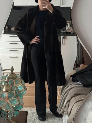 Mörkbrun lång fuskpäls kappa - Säljer en mörkbrun lång kappa i fuskpäls med breda ärmar och klassisk krage. Kappan har en rak och rymlig passform som ger en cool och elegant look. Perfekt för dig som vill ha något varmt och stilrent under kalla dagar.