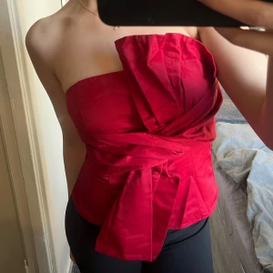 Röd axelbandslös topp med knyt - Snygg röd axelbandslös topp med en stor knytdetalj framtill. Perfekt för en kväll ute eller en speciell tillställning. Toppen har en figurnära passform och en dragkedja i ryggen för enkel på- och avtagning.