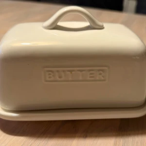 Smörburk i vitt porslin - Säljer ett klassiskt smörfat från märket JSHKY. Det är tillverkat i rejält, blyfritt porslin som känns både lyxigt och hållbart.  Passar perfekt för 500g smör.   Locket har ett praktiskt handtag och sitter stadigt på brickan utan att vingla. Fungerar lika bra i kylen som framme på köksbänken.  Enkel och stilren design som passar alla hem!