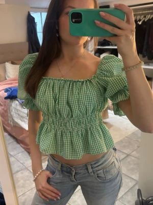 Grön rutig blus med puffärmar - Superfin grön och vit rutig blus med puffärmar och smock i midjan. Toppen är croppad och har en söt volang nertill. Perfekt för vår och sommar, och ger en fräsch och trendig look.
