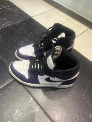 Nike Air Jordan 1 Court Purple High - Snygga Nike Air Jordan 1 sneakers i färgerna vitt, svart och lila. Skorna har högt skaft, svart swoosh och klassisk AJ1-design med perforerad tåbox och svarta skosnören. Materialet är en mix av läder och textil för en ikonisk look.