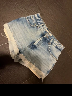 Ljusa jeansshorts från NA-KD - Säljer ett par ljusblå jeansshorts från NA-KD med hög midja och råa, fransiga kanter. Shortsen har klassisk femficksdesign och stängs med fyra silverfärgade knappar framtill. Perfekta för sommaren och riktigt snygga till sneakers eller sandaler.