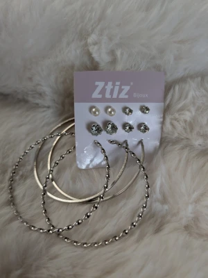 Örhängeset från Ztiz med pärlor och hoops - Snyggt örhängeset från Ztiz med två par stora silvriga hoops, ett par med tvinnad design och ett par släta. Inkluderar även tre par studs: ett par med vita pärlor och två par med glittrande stenar i olika storlekar. Perfekt för att mixa och matcha till olika looks.