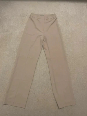 Beige kostymbyxa från H&M - Snygga beige kostymbyxor från H&M med rak passform och hög midja. Byxorna har klassisk design med dold knäppning och är tillverkade i ett mjukt, vävt material som ger en stilren look. Perfekta för dig som gillar minimalistisk och clean stil.