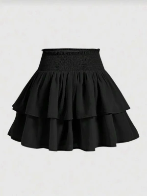 Svart volangkjol med resårmidja storlek M - Trendig svart kortkjol med dubbla volanglager och bred smockresår i midjan. Kjolen har en luftig och flowy känsla, perfekt för att styla med crop tops eller stickade tröjor. Enkel att matcha och ger en cool vibe till din outfit.