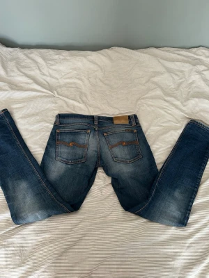 Blå jeans från Nudie Jeans - Säljer ett par klassiska blå jeans från Nudie Jeans med snygg slitning på framsidan och bakfickor med den ikoniska vågstickningen. Jeansen har normal midja, raka ben och är gjorda i mjuk denim. Perfekta för en avslappnad och trendig look. Hör av er vid frågor, pris är diskuterbart!🙂👍
