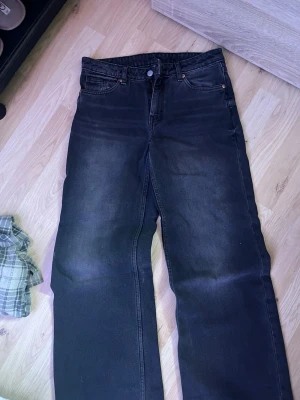Svarta raka jeans med tvättad look - Säljer ett par svarta jeans med rak passform och snyggt tvättad finish. Klassisk femficksmodell med knappgylf och bälteshällor. Jeansen har en avslappnad vibe och passar perfekt till en streetwear-outfit.