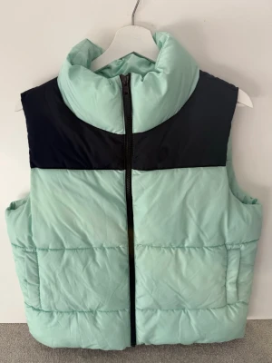 Mintgrön och svart dunväst - Snygg mintgrön dunväst med svart axelparti och hög krage. Västen har dragkedja framtill och två sidofickor. Perfekt för lager-på-lager och ger en cool streetwear-vibe. Materialet är quiltat och ger en puffig look.