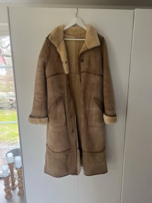 Lång brun fårskinnskappa - Säljer en lång brun fårskinnskappa med mjukt, ljust foder och bred krage. Kappan har knappar framtill, uppvikta ärmslut och två snedställda fickor. Perfekt för kalla dagar och ger en riktigt vintage och cozy vibe.