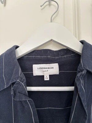 Mörkblå randig skjorta Lindbergh - Snygg mörkblå skjorta från Lindbergh med vita vertikala ränder och korta ärmar. Skjortan har klassisk krage och knäppning framtill. Tillverkad i ett lätt och luftigt material, perfekt för varmare dagar.