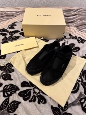 Svarta Axel Arigato sneakers - Stilrena svarta sneakers från Axel Arigato i mocka med minimalistisk design. Låg modell med svart sula och diskret logga på sidan. Perfekta för dig som gillar clean och modern stil. Skorna kommer med originalkartong och dustbag.