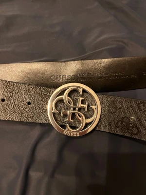 Svart bälte med silverdetaljer från Guess - Snyggt svart bälte från Guess med präglat logomönster och en stor rund silverspänne med Guess-logga. Bältet har en exklusiv vibe och passar perfekt till jeans eller byxor för att lyfta din outfit.