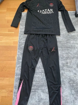 PSG träningsset svart Jordan x Nike - Säljer ett svart PSG träningsset med långärmad tröja och byxor. Setet har diskreta mönster, rosa detaljer på benen och PSG-logga samt Jordan-logga. Materialet är mjukt och stretchigt, perfekt för träning eller chill. Qatar Airways sponsortryck på bröstet.