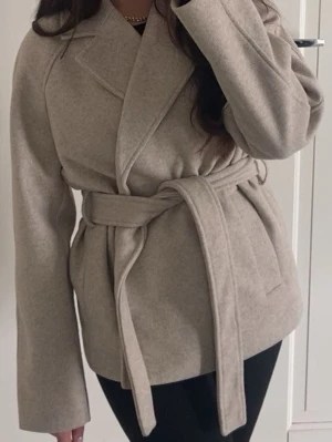 Beige kappa med knytskärp - Stilren beige kappa med bred krage och knytskärp i midjan. Kappan har en rak passform och är tillverkad i ett mjukt, ull-liknande material. Perfekt för dig som gillar minimalistisk och trendig stil. Nypris 700kr!!