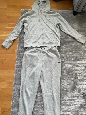 Grått tracksuit -set från Lacoste - Mysigt grått loungewear-set från Lacoste med zip-hoodie och matchande byxor. Hoodien har dragsko, fickor och dragkedja framtill. Byxorna har resår i midjan och diskret Lacoste-logga på både tröja och byxa. Perfekt för chill dagar hemma.
