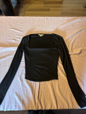 Svart långärmad topp med fyrkantig ringning - Snygg svart långärmad topp med fyrkantig ringning och tight passform. Perfekt att styla med jeans eller kjol för en trendig look. Tillverkad i mjukt och stretchigt material som sitter skönt på kroppen.