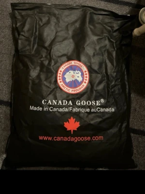 Svart dunväst från Canada Goose - Säljer en svart dunväst från Canada Goose med hög krage och tryckknappar framtill. Västen har två stora fickor och den klassiska Canada Goose-loggan på bröstet. Perfekt för kalla dagar när du vill hålla värmen och ändå se stilren ut. Kommer med påse 