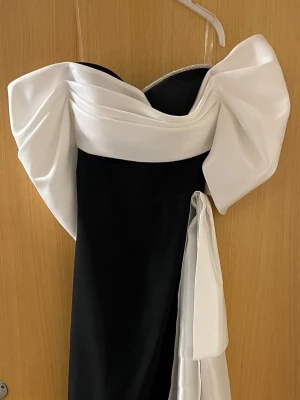 Svart och vit axelbandslös klänning - Snygg svart och vit axelbandslös klänning med stor rosett baktill och draperad överdel. Klänningen har en elegant, lång siluett och är dekorerad med en glittrig kant upptill. Perfekt för dig som vill sticka ut på festen.