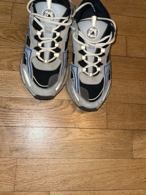 Grå och blå sneakers från Axel Arigato - Chunky sneakers från Axel Arigato i grått, svart och blått med vita detaljer. Skorna har en sportig design med tjock sula, mesh och mocka-paneler samt vita snören. Perfekta för dig som gillar streetwear och vill sticka ut med coola detaljer. Priset kan absolut diskuteras skorna finns inte längre på lager blivit sold out överallt så passa på 
