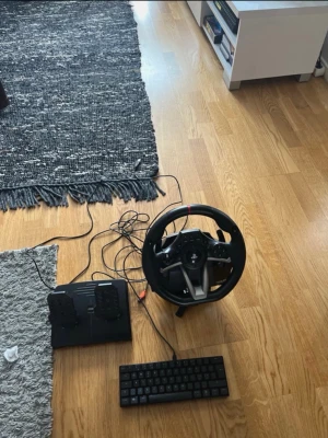 Hori ratt och pedaler till PS4/PC - Säljer ett svart racing rattset från Hori med tillhörande pedaler, perfekt för bilspel på PS4 eller PC. Ratten har flera knappar och en röd detalj upptill, och pedalerna är i plast med halkskydd. Allt kopplas in med sladdar för stabil anslutning.