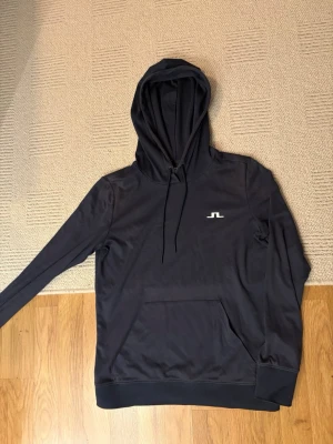 Svart hoodie från J.Lindeberg - Snygg mörkblå hoodie från J.Lindeberg med klassisk huva och dragsnören. Framtill finns en vit logga och en stor magficka. Tillverkad i mjukt och sportigt material som känns skönt mot huden. Perfekt för en avslappnad och stilren look.