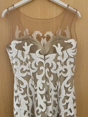 Beige klänning med vit spetsdetalj - Snygg beige klänning med transparent mesh upptill och dekorativa vita spetsapplikationer i ett unikt mönster. Klänningen har ärmlös design och figurnära passform, perfekt för dig som vill sticka ut. Ingen synlig dragkedja eller knappar.