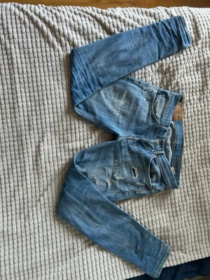 Dondup Jeans - Säljer ett par blå Dondup jeans med slitningar och hål på framsidan. Jeansen har fem fickor, normal midja och smal passform. Perfekta för en avslappnad och trendig look. Materialet är klassisk denim i bomull.