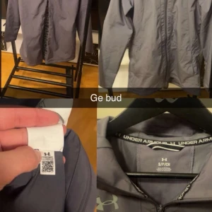 Grå vindjacka från Under Armour - Snygg grå vindjacka från Under Armour med dragkedja framtill och huva. Jackan har logga på bröstet och är tillverkad i ett lätt, vindtätt material som passar perfekt för blåsiga dagar. Enkel och stilren design som funkar till det mesta.