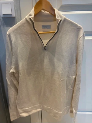 Beige halvzip tröja från Tommy Hilfiger - Snygg beige långärmad tröja från Tommy Hilfiger med halvzip och hög krage. Tröjan har en minimalistisk design och är tillverkad i mjukt material, perfekt för en avslappnad men stilren look.