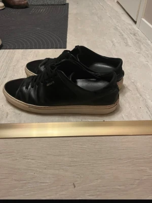 Svarta sneakers från Axel Arigato - Snygga svarta sneakers från Axel Arigato i läder med klassisk låg profil och vit sula. Diskret logga på sidan och stilren design som passar till det mesta. Skorna har svarta skosnören och en bekväm innersula.