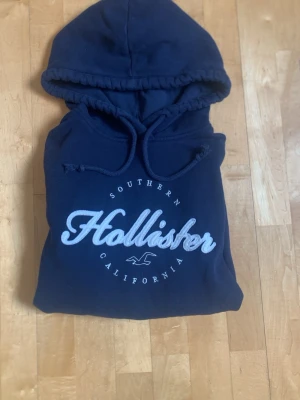Mörkblå hoodie från Hollister - Snygg mörkblå hoodie från Hollister med stor broderad logga och texten 'Southern California' på bröstet. Hoodien har dragsko i huvan och är tillverkad i mjukt material som känns skönt mot huden. Perfekt för en avslappnad och trendig look.