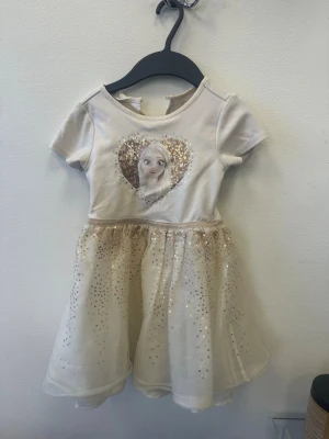 Klänning i ljusbeige  - Söt kort klänning i ljusbeige med glittrigt hjärta framtill och ett lager av skimrande tyll över kjolen. Klänningen har korta ärmar och rund halsringning. Perfekt för speciella tillfällen när du vill glänsa lite extra.