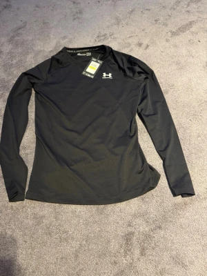 Svart långärmad träningströja UA - Snygg svart långärmad träningströja från Under Armour med vit logga på bröstet. Tillverkad i funktionsmaterialet HeatGear som andas och sitter tight för bästa komfort vid träning. Perfekt för gymmet eller löprundan.
