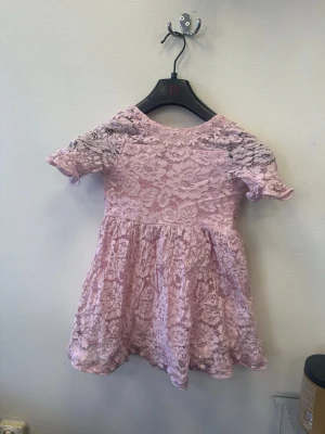 Rosa spetsklänning med rosett - Söt rosa kortklänning i spets med korta ärmar och en stor sidenrosett i midjan baktill. Klänningen har ett blommigt spetsmönster och en öppen detalj i ryggen. Perfekt för speciella tillfällen när du vill känna dig extra fin.