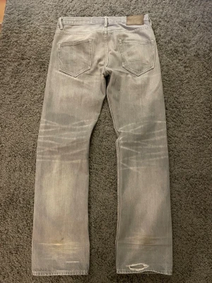 Grå raka jeans från all saints  - Säljer ett par gråa  jeans med raka ben och klassisk femficksmodell. Jeansen har slitningar och en ljus tvätt som ger en cool vintagekänsla. Tillverkade i bomull med en skön passform. Perfekta för en avslappnad streetstil. Nypris runt 1500
