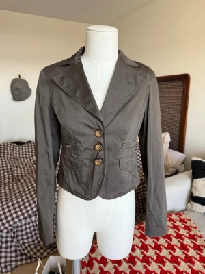 Grå kavaj / blazer  - Grå/grön kavaj med figurnära passform och knappar framtill. 100 % bomull. Sparsamt använd, fint skick. Liten i storleken. Storlek L, men passar mig fint som är en storlek S. Köpt second hand. Tillverkad i Italien enligt materiallapp. 