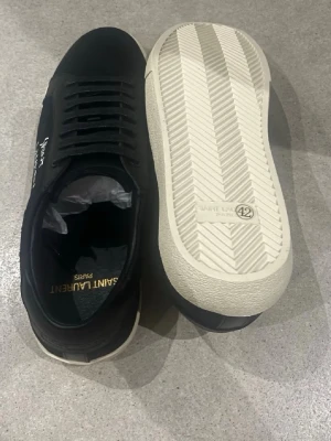Svarta Saint Laurent sneakers - PRIS KAN DISKUTERAS!!! Svarta sneakers från Saint Laurent med vit sula och guldfärgad logga på insidan. Skorna har lågt skaft, klassisk snörning och är tillverkade i läder. Snygg och stilren design med diskret Saint Laurent-tryck på sidan.