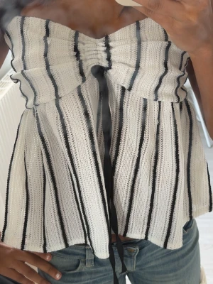 Randig bandeau topp med snörning - Supersnygg bandeau topp i vitt med svarta vertikala ränder. Toppen har en söt snörningsdetalj framtill och är tillverkad i ett stickat material som ger en luftig och bekväm känsla. Perfekt för varma dagar och enkel att styla med jeans eller kjol.