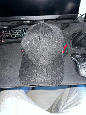 Svart Gucci keps med GG-mönster - Snygg svart keps från Gucci med klassiskt GG-monogram över hela kepsen och en grön-röd rand på sidan. Kepsen har böjd skärm och justerbar rem bak. Materialet ser ut att vara canvas med detaljer i tyg. Perfekt för dig som vill ha en exklusiv och trendig look.