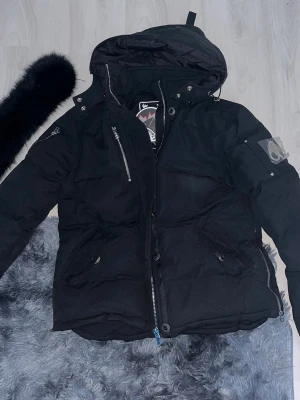 Svart pufferjacka från Moose Knuckles - Snygg svart pufferjacka från Moose Knuckles med stor huva och silverfärgade dragkedjor. Jackan har två framfickor, en ärmficka med metallogo och detaljer i metall. Perfekt för kalla dagar och riktigt street.