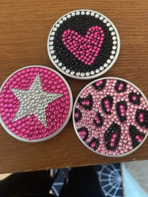 Y2K Sminkspeglar - Tre runda sminkspeglar med glittrande strass på ovansidan. En har ett rosa hjärta på svart botten, en har en silverstjärna på rosa botten och en har rosa och svart leopardmönster. Speglarna är kompakta och perfekta att ha i väskan. Material: metall och plast.