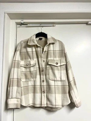 Beige och vit rutig overshirt - Overshirt i beige och vitt rutigt mönster med två stora bröstfickor och knappar framtill. Skjortkrage och avslappnad passform. Tillverkad i mjukt, tjockare tyg som ger en mysig känsla. Perfekt lager på lager-plagg.