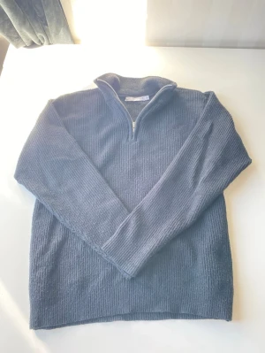 Svart stickad halvzip tröja - Svart stickad tröja med halvzip och hög krage. Tröjan har långa ärmar och är ribbstickad för en snygg och enkel look. Perfekt för lager på lager och passar till jeans eller chinos. Tröjan har också ett litet hål på högra armen vid armbågen lätt att fixa den 