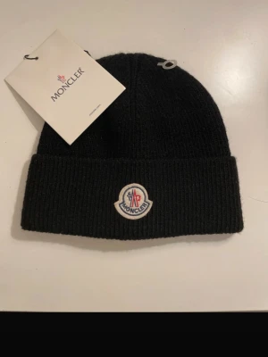 Svart Moncler mössa med logga - Snygg svart ribbstickad mössa från Moncler med uppvikt kant och klassisk Moncler-logga framtill. Tillverkad i mjukt material som håller dig varm och stilren under kalla dagar. Perfekt accessoar för dig som vill ha en clean och exklusiv look.