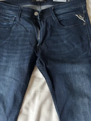 Mörkblå slim fit jeans från Replay - Snygga mörkblå slim fit jeans från Replay med klassisk femficksmodell och diskreta slitningar. Jeansen har dragkedjegylf och detaljerad Replay-logga vid fickan. Tillverkade i mjukt denimtyg som sitter skönt och följer kroppen.